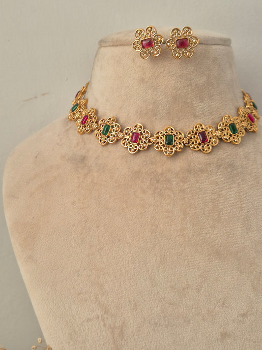 Traditional Indian antique gold-finish choker featuring connected floral motifs with alternating rectangular ruby-red and emerald-green gemstones, paired with matching floral stud earrings set with ruby-red stones