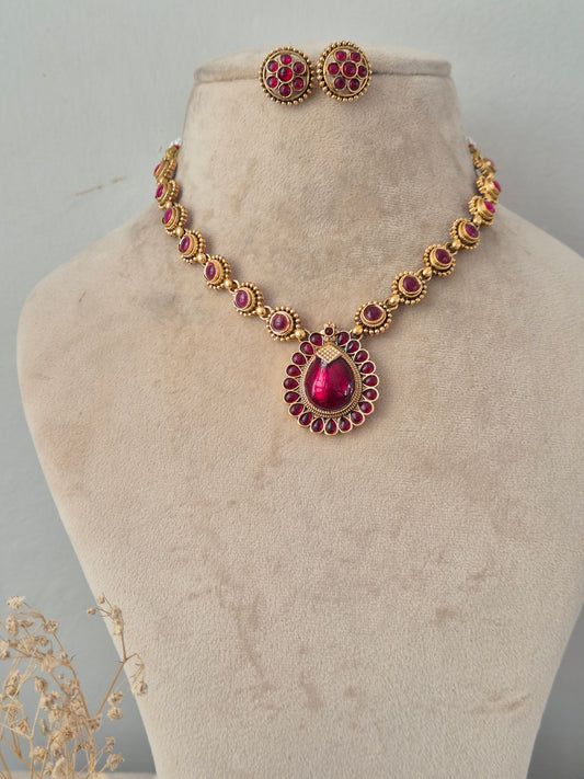 Traditional Indian antique gold necklace set featuring a chain studded with oval ruby-pink stones leading to a central teardrop ruby-pink pendant framed by smaller stones, paired with matching round floral stud earrings