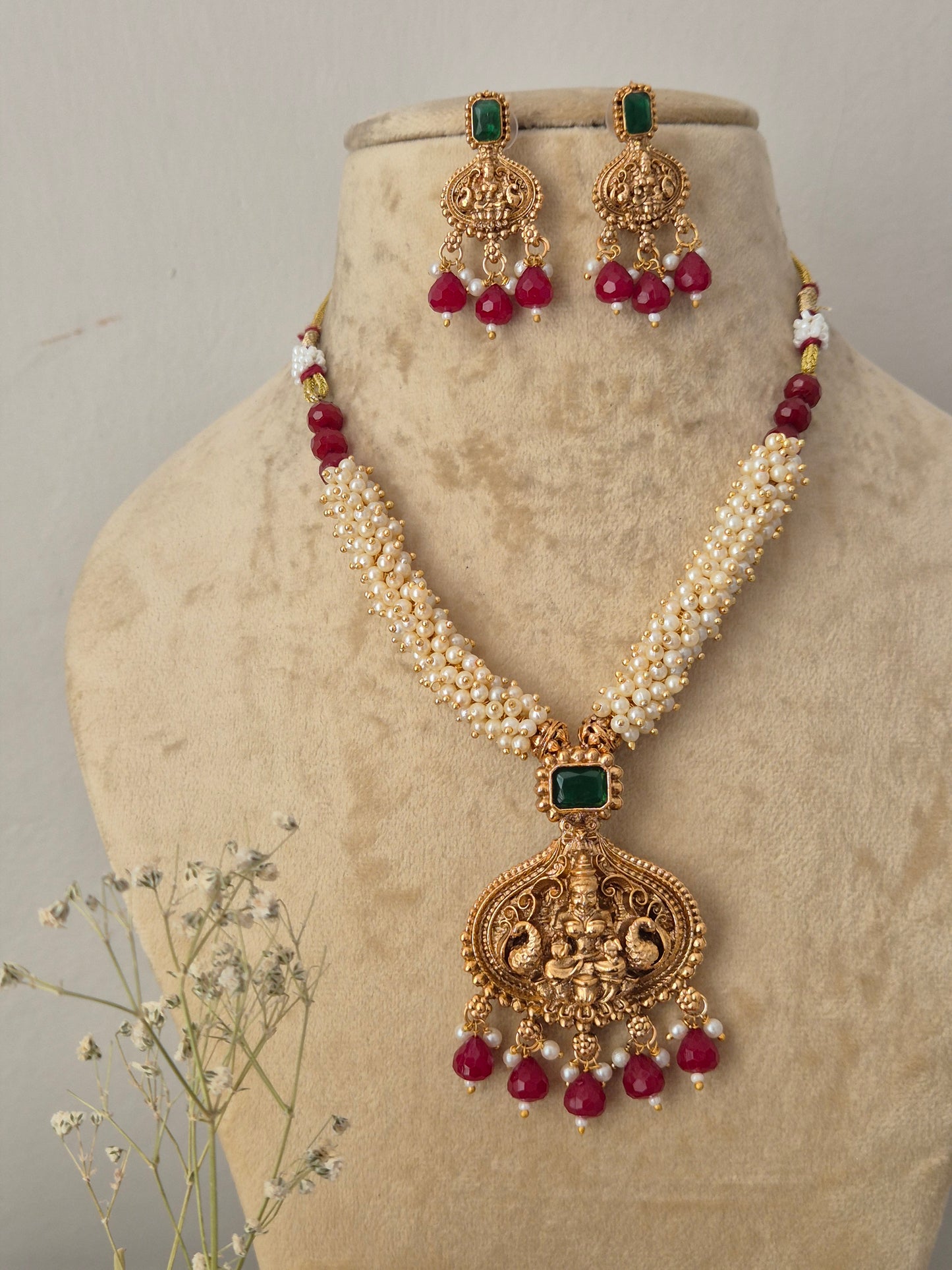 Gold-plated traditional Indian jewelry set featuring a necklace and matching earrings with white pearl-like beads, green rectangular stones, and red round beads, Naitri featuring an ethnic gold-plated temple pendant, multi-layered pearl chains, and matching statement drop earrings with red and green stones by naitri.in