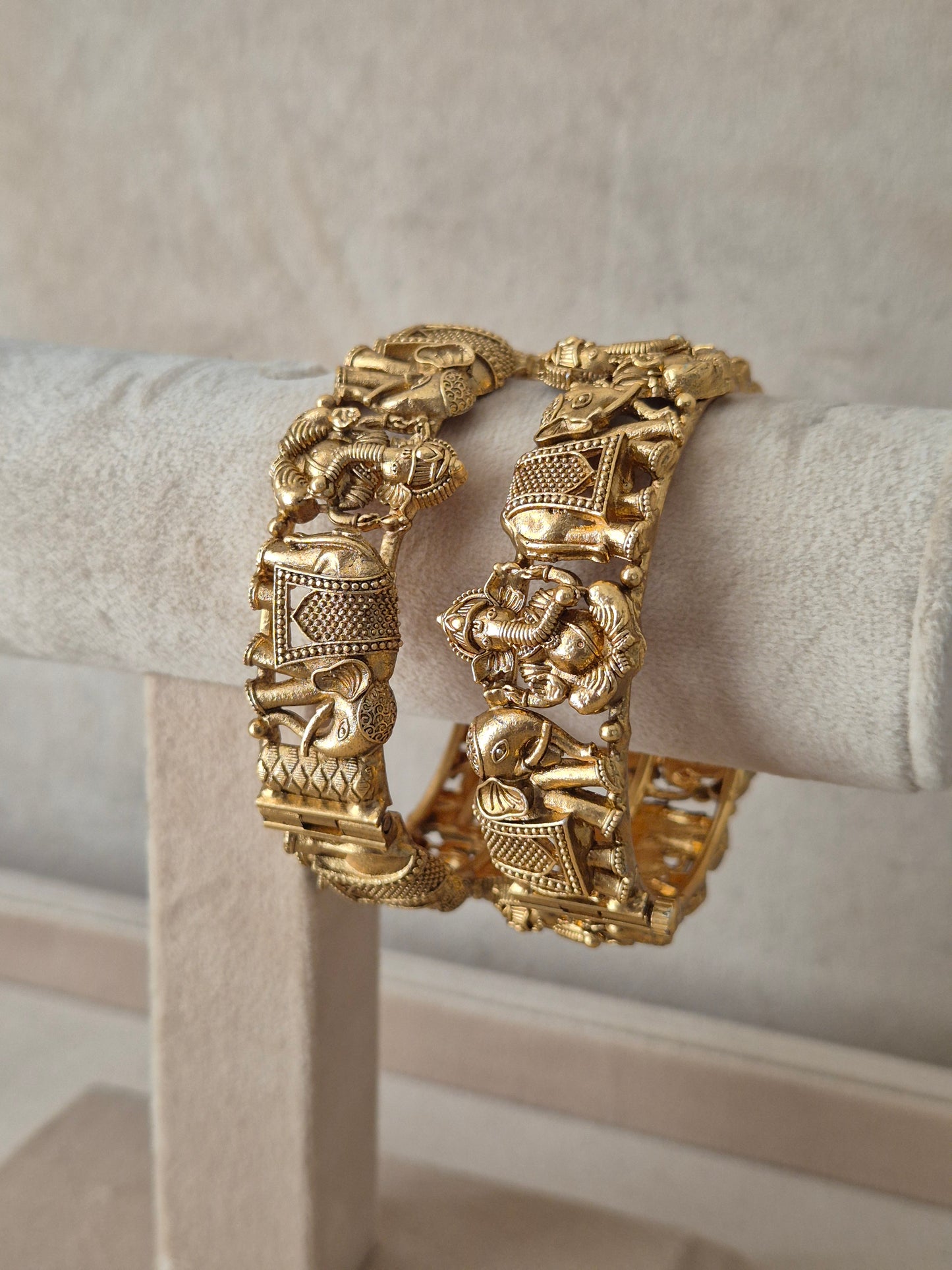 legant golden statement bangles with ethnic animal motif designs exhibited on a neutral luxury jewelry display next to small white flowers by naitriin