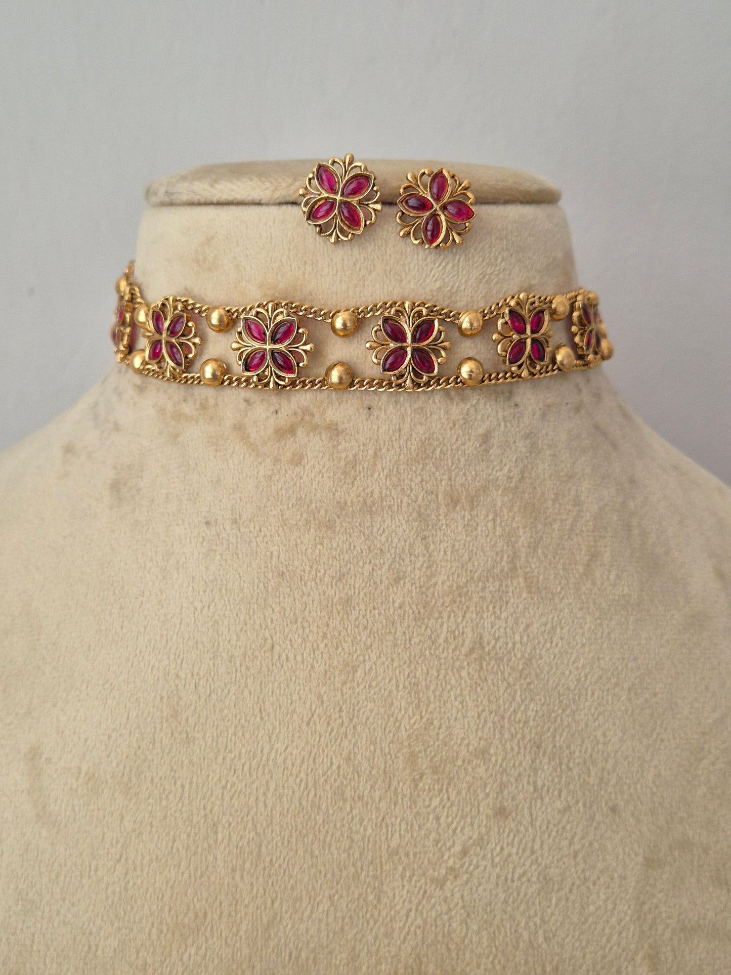 A gold-toned bracelet and matching stud earrings, jewelry features repeating floral motifs with deep pinkish-red centers and small gold bead accents by naitri.in