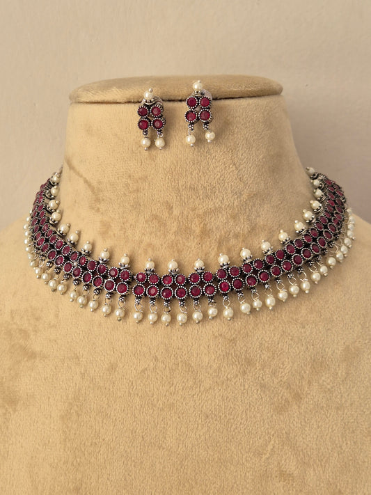 A traditional Indian jewelry set featuring a choker necklace and matching earrings, The set features silver-plated metal work, deep red stones, and white faux pearls by naitri.in