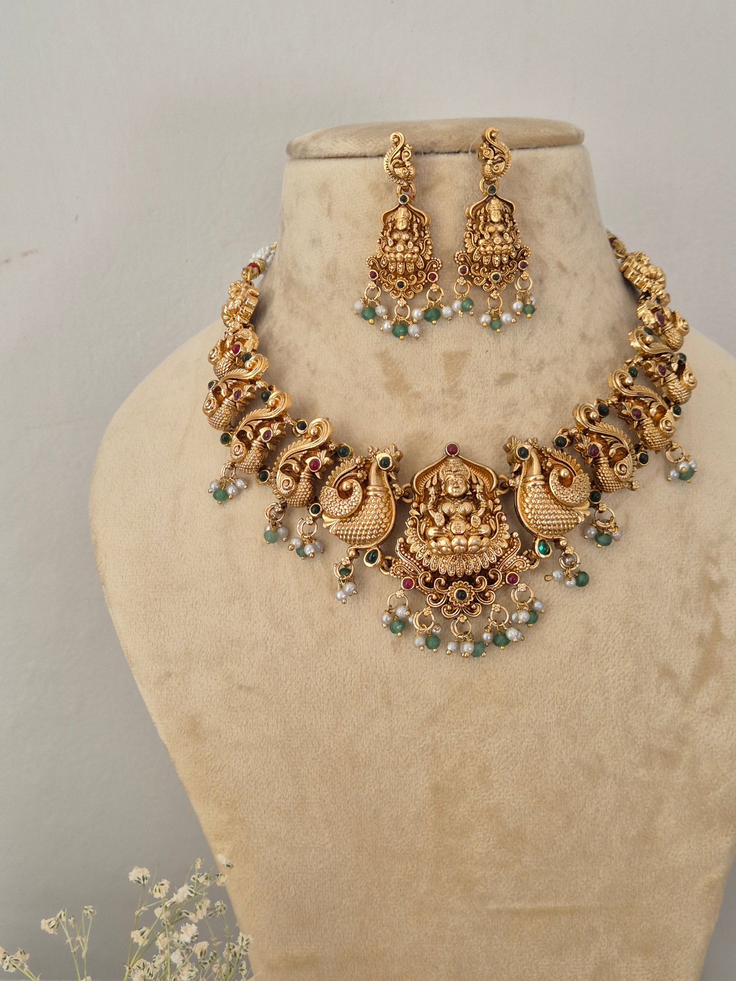 Gold-plated traditional Indian jewelry set featuring a choker necklace and matching drop earrings, decorated with peacocks motifs, green and red stone accents, and white pearl beads, featuring an antique gold-plated ethnic choker and matching temple jewelry drop earrings for festive wear by naitri.in