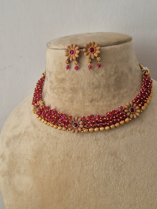 Modern Indian ethnic imitation jewelry set including a golden brass alloy choker with vibrant pink-red gemstones in a layered strand design, shown with matching floral-themed earrings by naitri.in