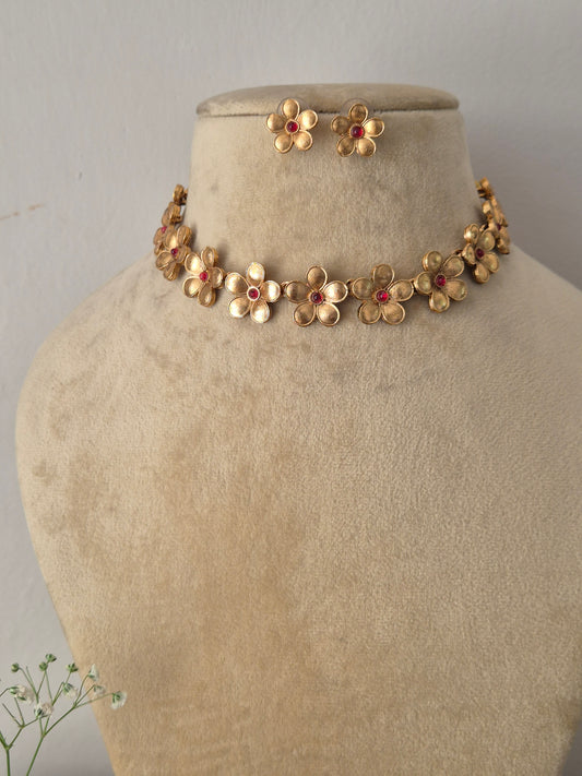 Modern Indian ethnic imitation jewelry set including a golden brass alloy choker necklace crafted with delicate floral motifs and pinkish-red stone centers, shown with matching flower earrings by naitri.in
