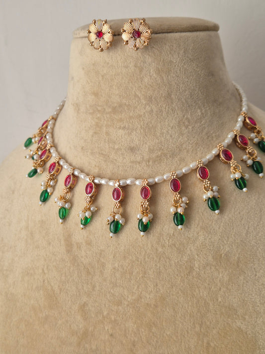 Traditional Indian antique-finish brass jewelry set showcasing a dainty pearl collar layout, vibrant pinkish-red and green gemstone charms, and coordinating floral-stamped stud earrings from the Naitri collection by naitri.in