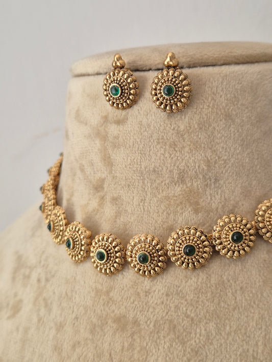 Antique gold-toned traditional choker necklace and matching stud earrings. Both the choker and matching circular earrings feature intricate, textured sunflower-like metallic patterns surrounding a central deep green gemstone by naitri.in