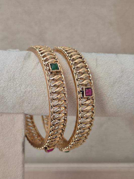A pair of gold-plated bangles with a textured chain-like pattern, featuring small square green and pink gemstone accents, by naitri.in