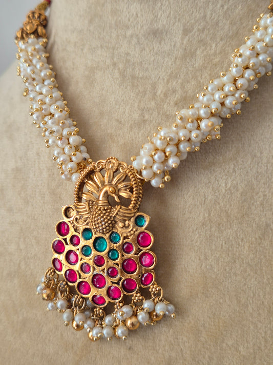 Traditional Indian gold-polished brass necklace set showcasing an intricate antique filigree pendant with ruby-red and emerald-green gemstones, suspended from a woven pearl strand by naitri.in