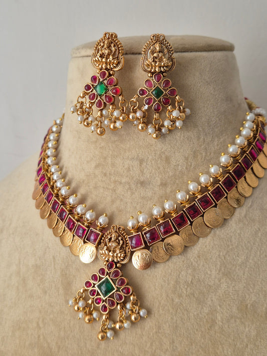 Traditional Indian temple jewelry set in polished brass, showcasing an antique finish pendant with red and green gemstone accents, a layered pearl band, and a classic coin fringe by naitri.in