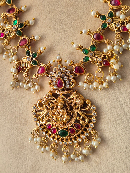An ornate gold-toned traditional jewelry piece featuring a central deity motif, embellished with red and green teardrop-shaped stones, round clear stones, and small white pearl-like beads, by naitri.in