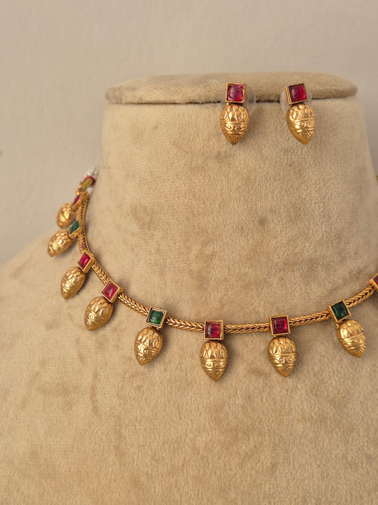 Gold jewelry set featuring a necklace and matching earrings with textured gold leaf pomegranate-shaped charms and square red-green gemstone accents, by naitri.in