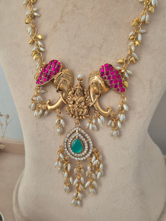 gold temple necklace featuring magenta stone-encrusted elephant motifs, a green teardrop center pendant with dangling pearls, Gold temple necklace set with pink elephant accents, a green stone pendant, and pearl drop details by naitri.in