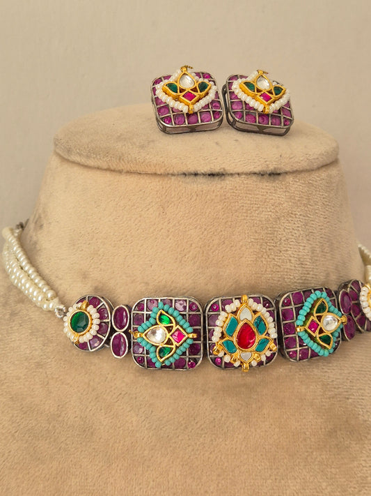 A jewelry set featuring a statement necklace and matching earrings. The jewelry has silver-toned metal settings with purple gemstone borders, central red and turquoise stones, and white pearl-like accents by naitri.in