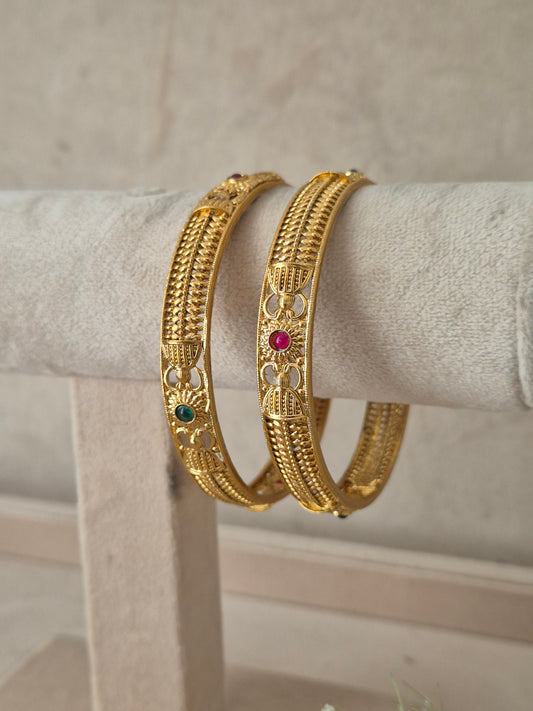A pair of gold-plated Indian bangles with intricate filigree work and small red and green stone accents, by naitri.in