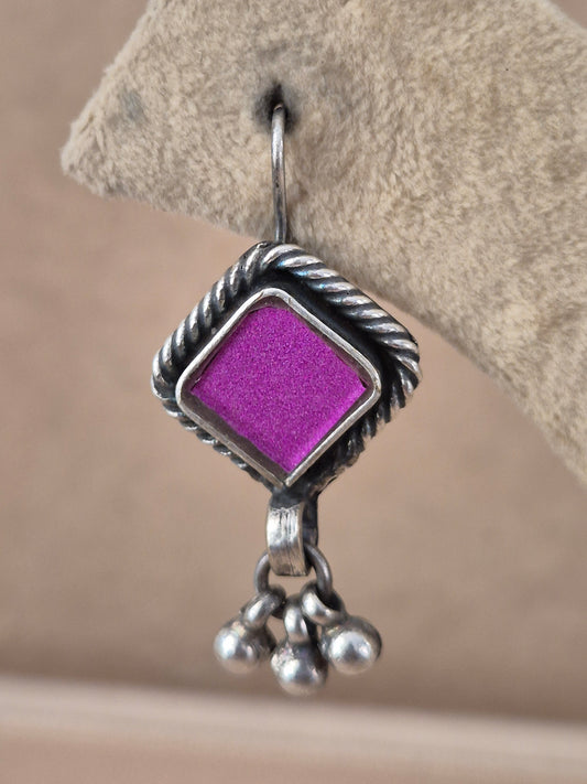 A close-up of a silver dangle earring featuring a textured, twisted border and a bright purple square center, hanging from a silver fishhook by naitri.in