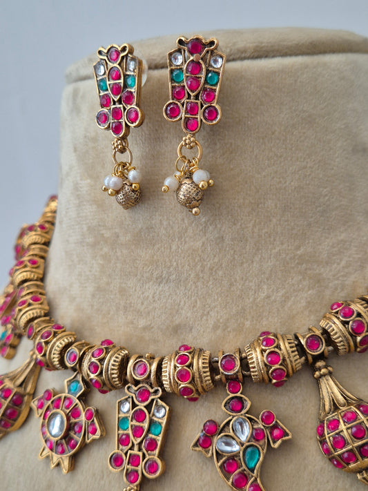 Traditional Indian imitation jewelry set showing a brass alloy gold necklace studded with pink and green gemstones, tiny pearls, and matching ethnic earrings by naitri.in