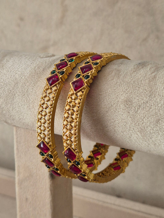 A pair of gold-plated bangle bracelets decorated with rectangular red gemstones and small green stone accents, by naitri.in