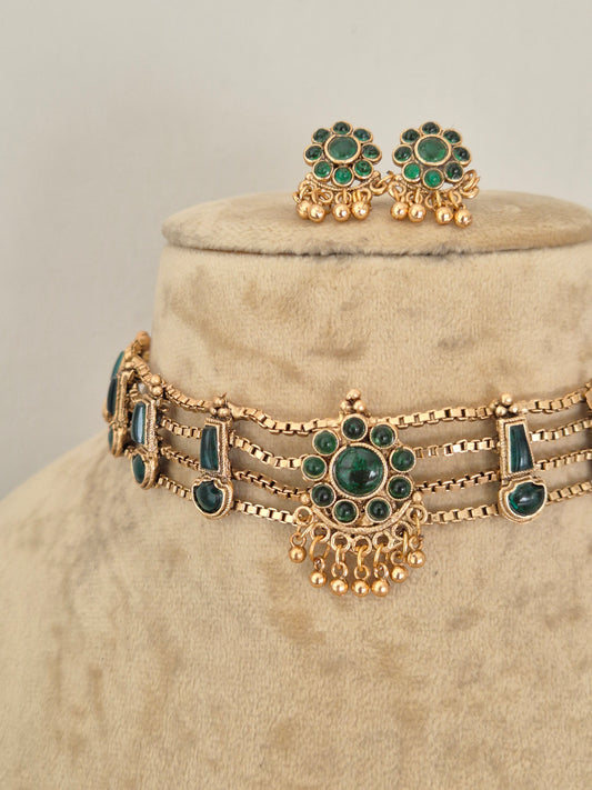 Antique gold-toned choker necklace showcasing an intricate metallic floral band, vibrant green stone clusters, and delicate hanging gold bead drops, shown with coordinating round stud earrings by naitri.in