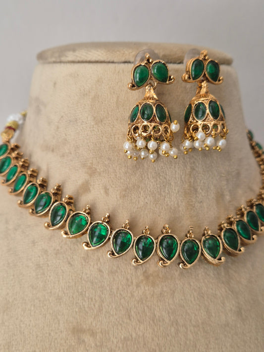 Gold-toned jewelry set with emerald green teardrop stones and small white pearl accents. The set includes a necklace and matching earrings. The choker features a row of vibrant oval-cut green gemstones bordered by tiny gold accents, paired with matching bell-shaped jhumka-style earrings embellished with green stones and hanging white pearl droplets by naitri.in