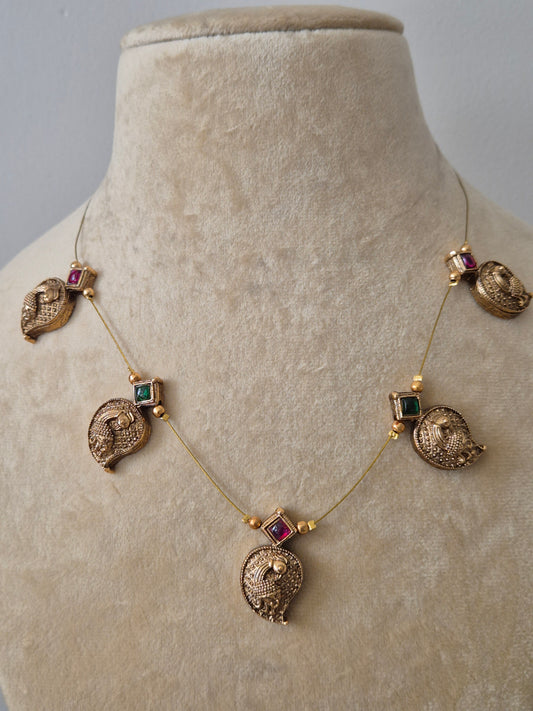 Handcrafted peacock pendant necklace with an invisible chain design, featuring intricate gold-polished brass metalwork and vibrant ruby-red stone details by naitri.in