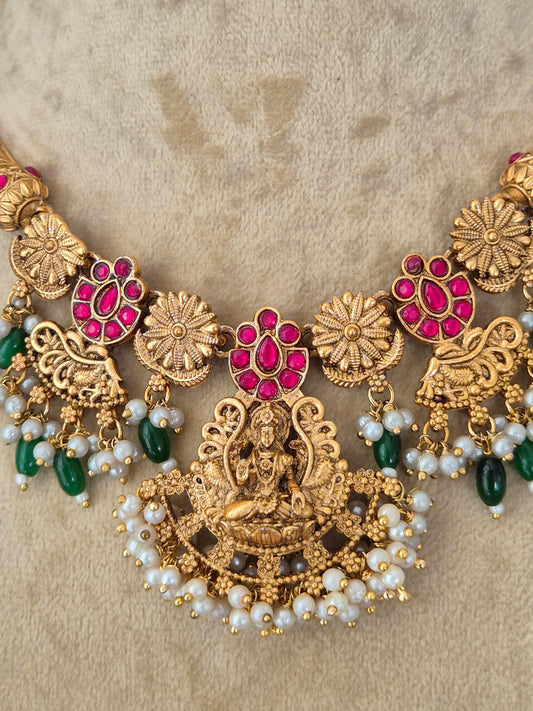 An ornate gold-toned traditional necklace with red gemstone floral motifs, white pearl bead strands, and green bead accents, necklace is designed with intricate floral metalwork, red enamel accents, green gemstones, and a fringe of hanging white pearl-like beads by naitri.in
