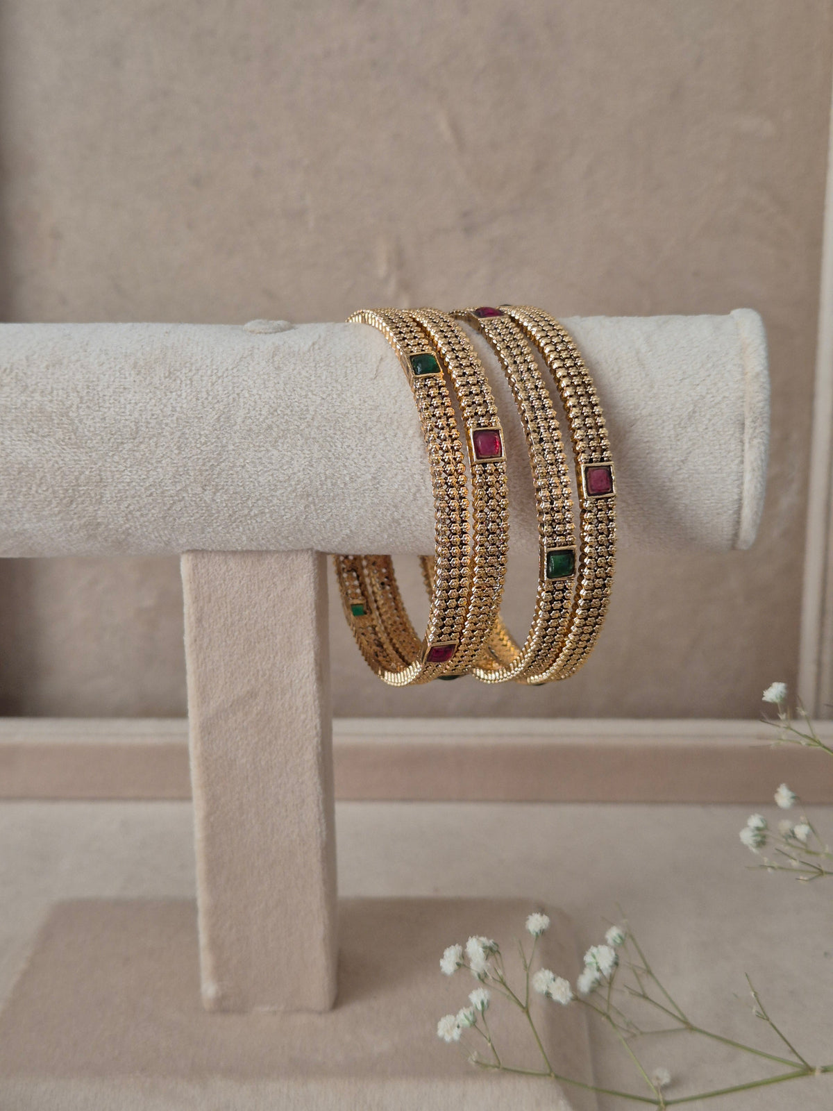 A stack of four Naitri Savya gold bangles showcasing detailed traditional craftsmanship, featuring vibrant pinkish-red and green accent stones, by naitri.in