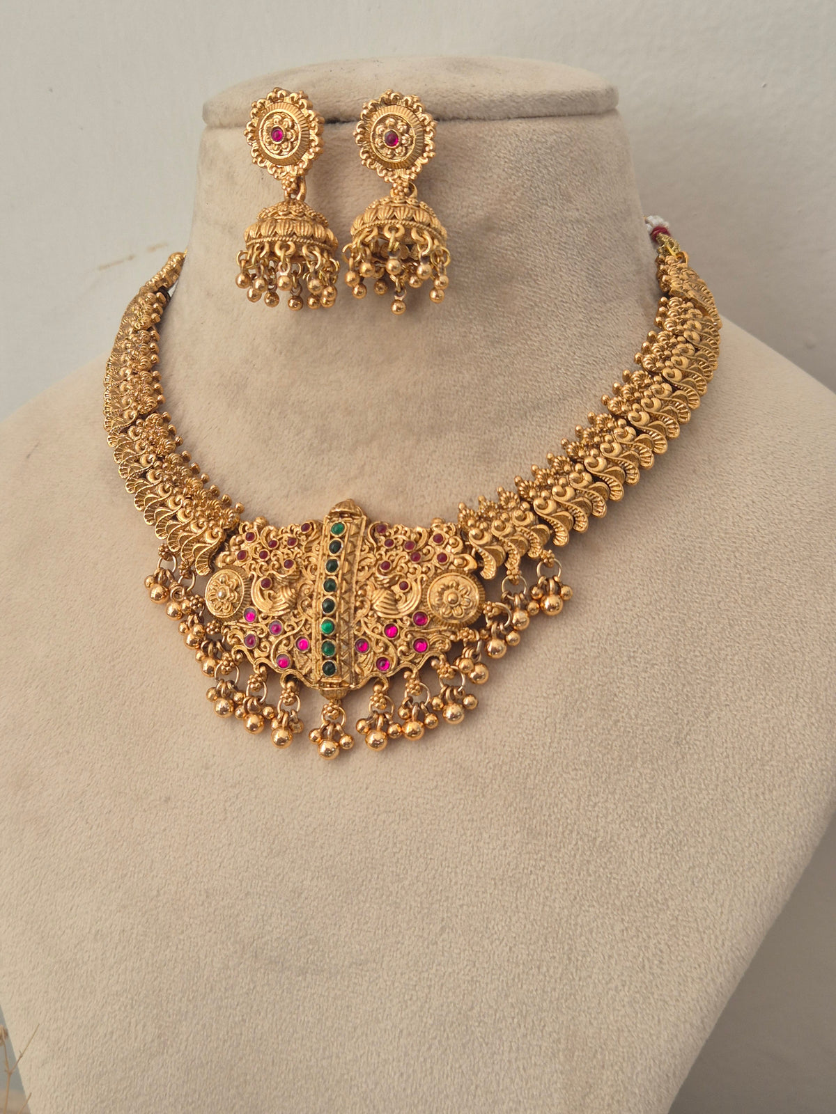 Gold-toned traditional Indian jewelry set featuring a wide collar necklace and matching jhumka earrings, decorated with gold coins, filigree, and red and green stones, by naitri.in