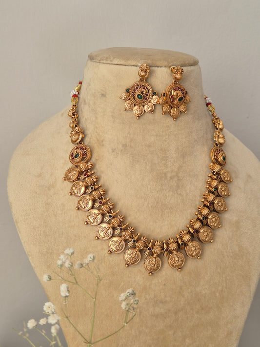 Traditional Indian gold coin necklace set with an antique finish, intricately carved floral and deity-style motifs, and small ruby-red and emerald-green stone embellishments by naitri.in