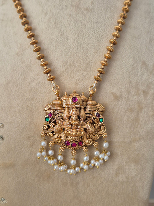 An ornate gold-toned traditional pendant necklace with a deity motif, accented with red and green stones and dangling pearl beads by naitri.in