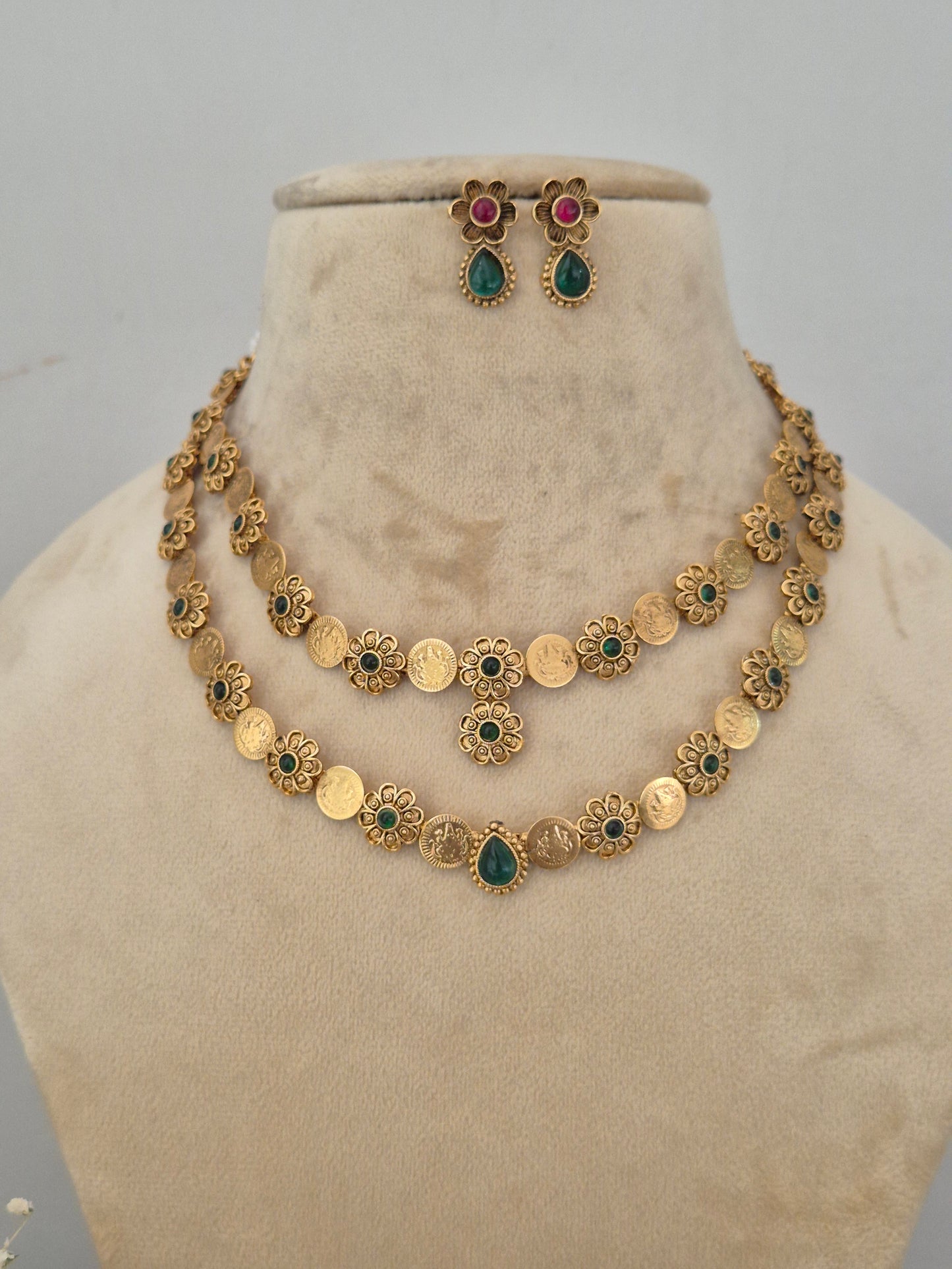 A gold-toned traditional necklace and matching earrings. The necklace features round gold coin-like charms, floral filigree details, and small green and red stone accents. by naitri.in