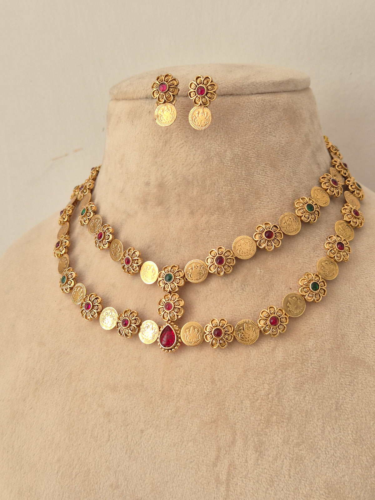 Yamya gold-toned double-layer coin necklace with red and green stones, styled with matching floral coin earrings on a mannequin bust.
