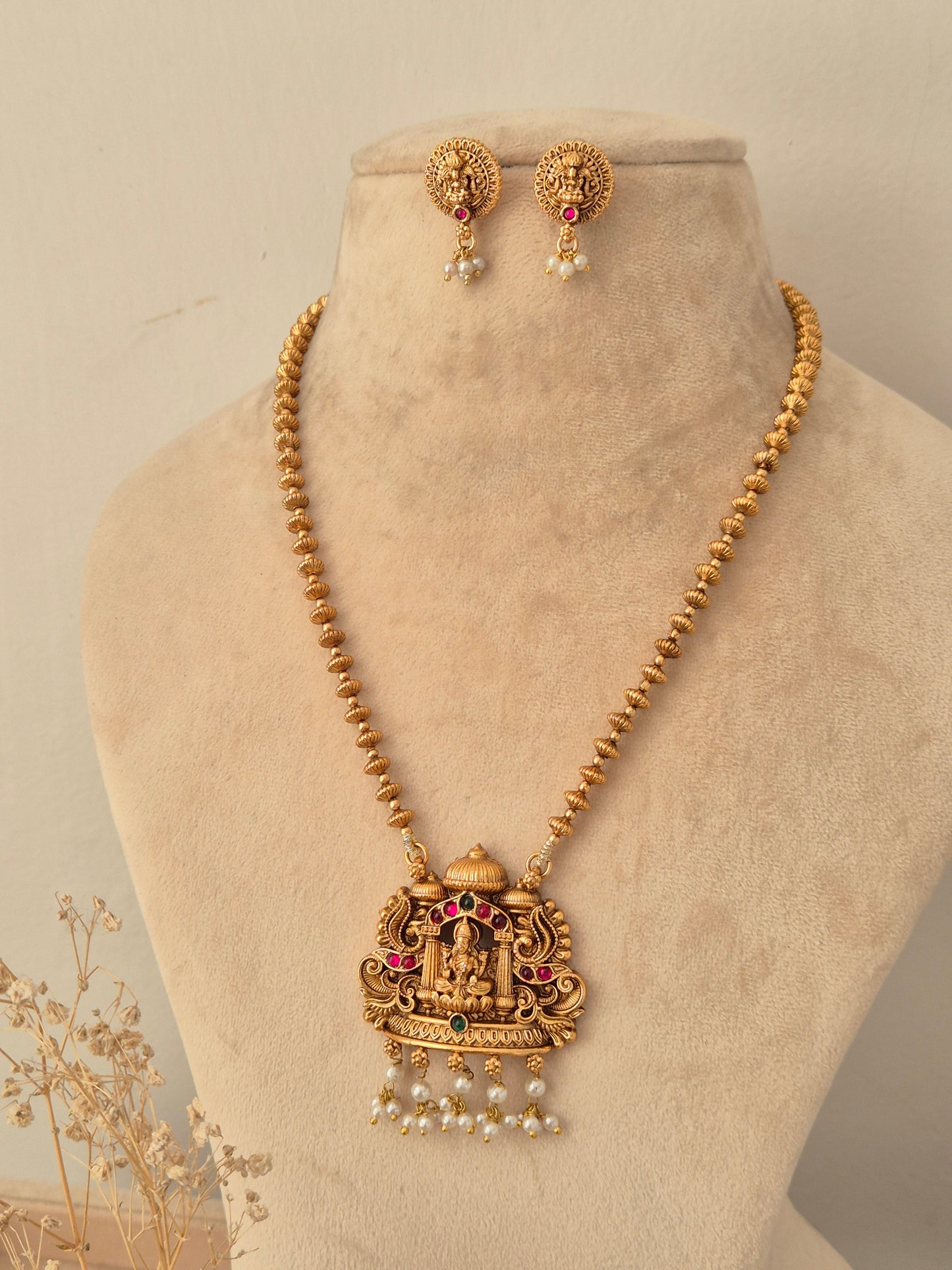 Yamira long temple necklace set with Goddess Laxmi pendant, red and green stones, pearl drops, and matching stud earrings styled on a mannequin bust.