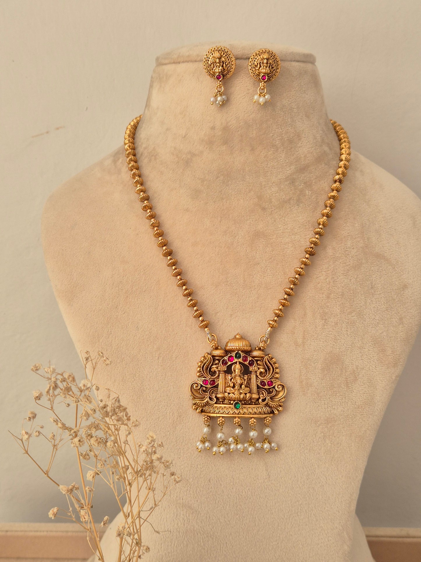 Traditional Indian long gold-toned beaded chain necklace featuring a detailed Goddess Laxmi temple pendant with pink, red, and green stone embellishments and hanging white pearl drops, styled with matching round stud earrings 