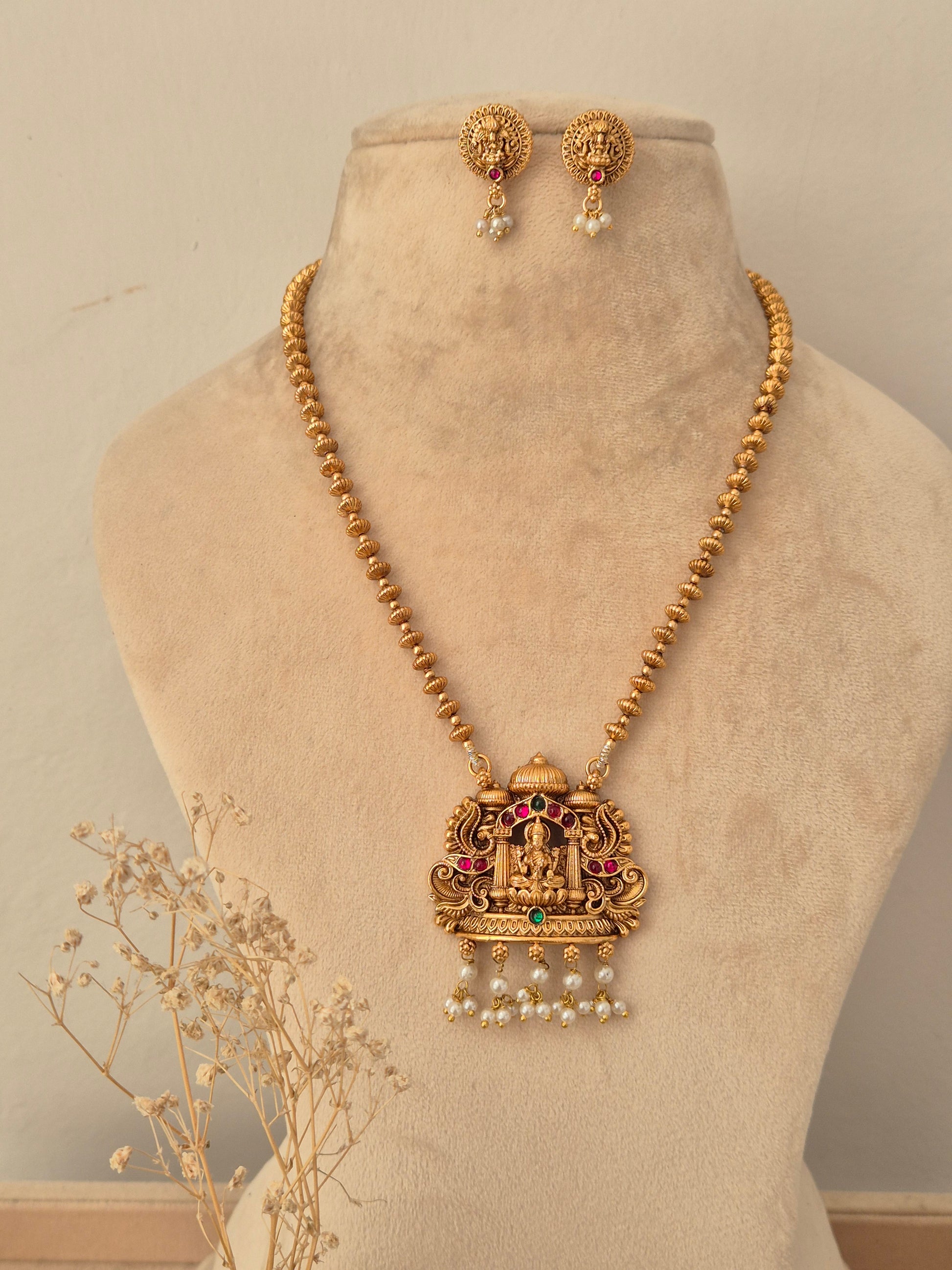 Traditional Indian long gold-toned beaded chain necklace featuring a detailed Goddess Laxmi temple pendant with pink, red, and green stone embellishments and hanging white pearl drops, styled with matching round stud earrings 