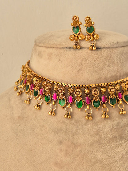 Gold-finish choker necklace adorned with teardrop-shaped pink and green stones, small dangling beads, and matching stud earrings shown on a beige jewelry display