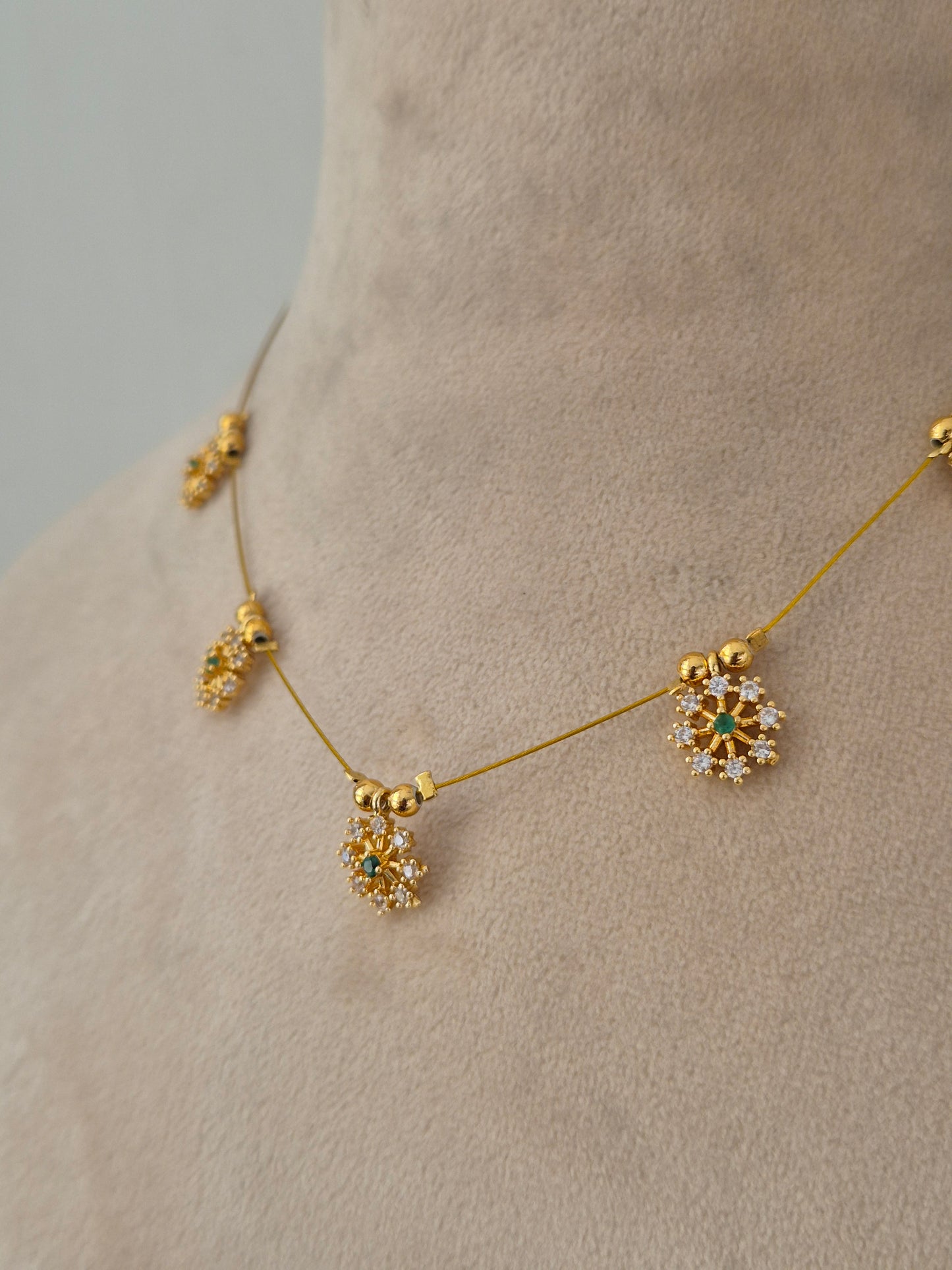 Delicate gold-toned necklace designed with a minimal transparent wire appearance, holding five sparkling floral starburst charms set with clear and green stones