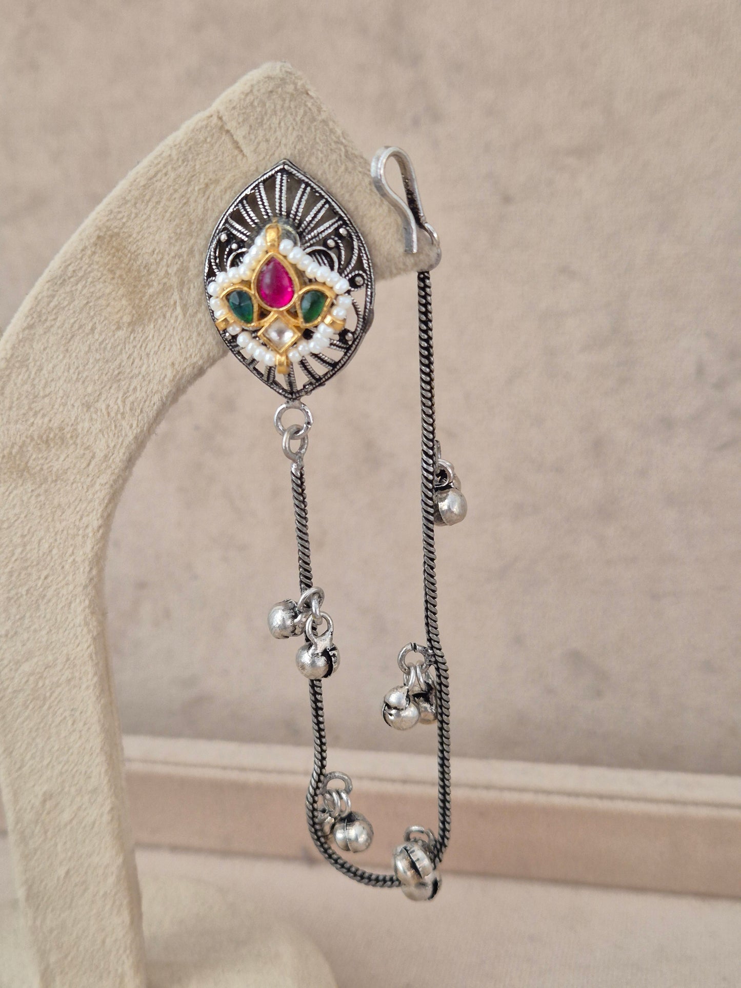 Pair of silver-finish earrings with diamond-shaped stud tops set with colorful pink and green stones, styled with multiple long hanging chains and small bell charms on a beige display stand