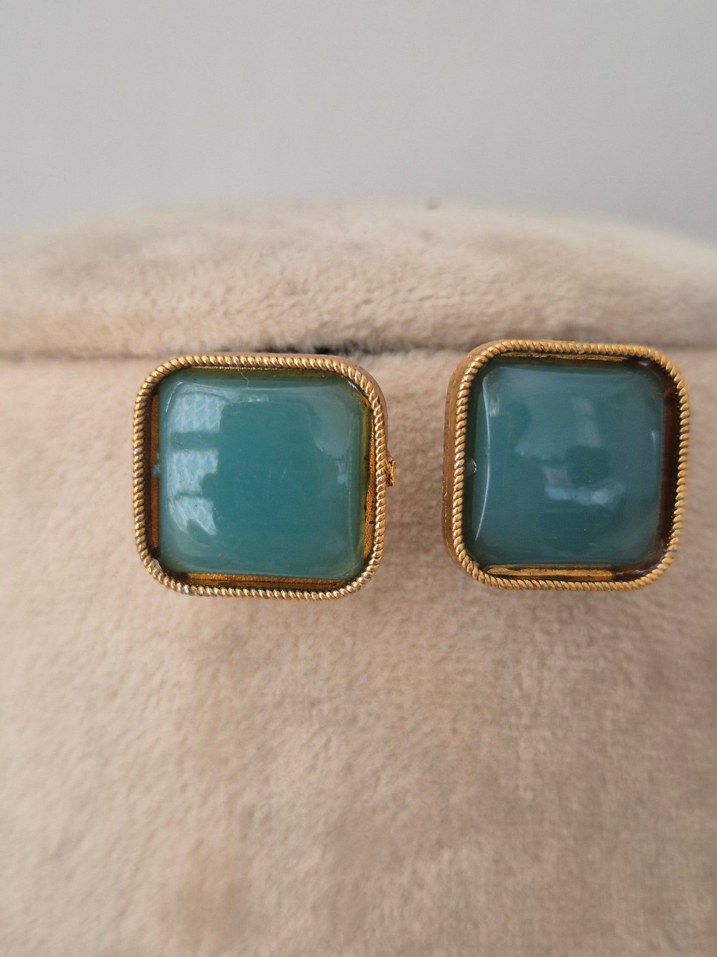 Close-up of matching square teal stud earrings with twisted gold-toned borders displayed on a neutral velvet display surface