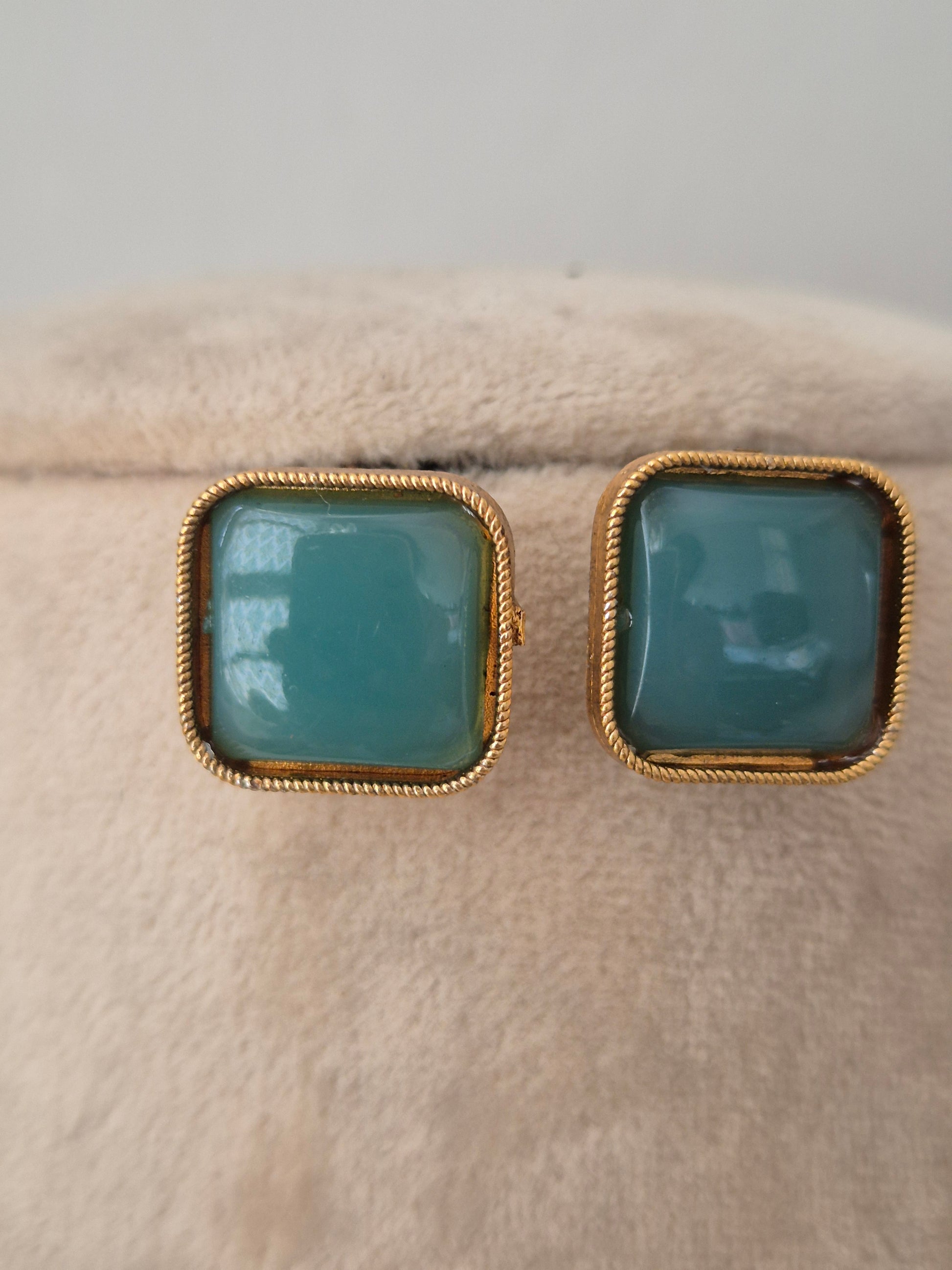 Close-up of matching square teal stud earrings with twisted gold-toned borders displayed on a neutral velvet display surface