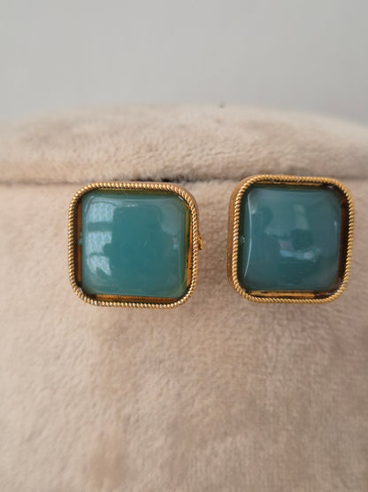 Close-up of matching square teal stud earrings with twisted gold-toned borders displayed on a neutral velvet display surface