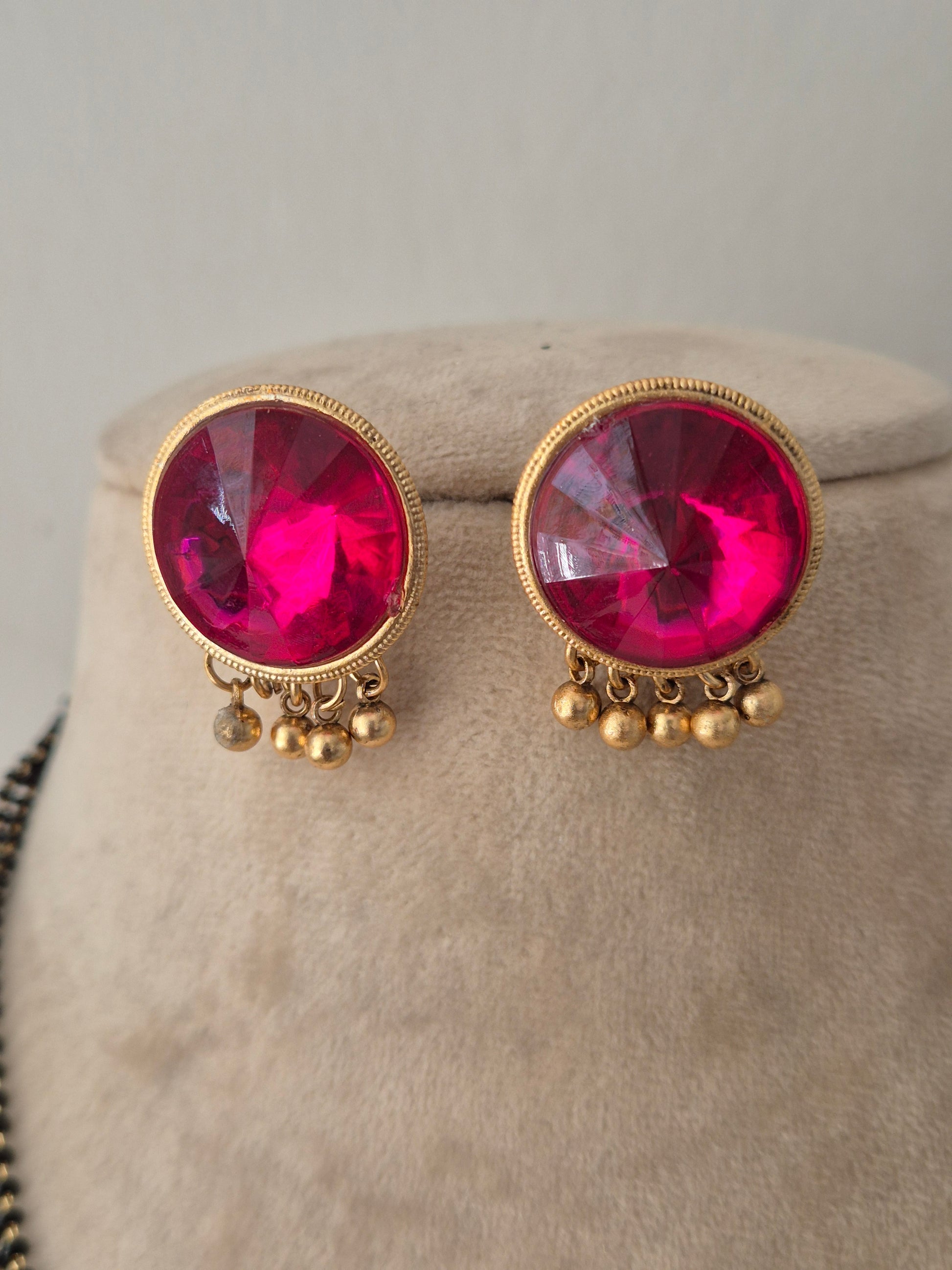 Pranati Mangalsutra Set – Pair of round magenta pink gemstone stud earrings with gold beaded drop fringe accents, displayed on a neutral fabric bust