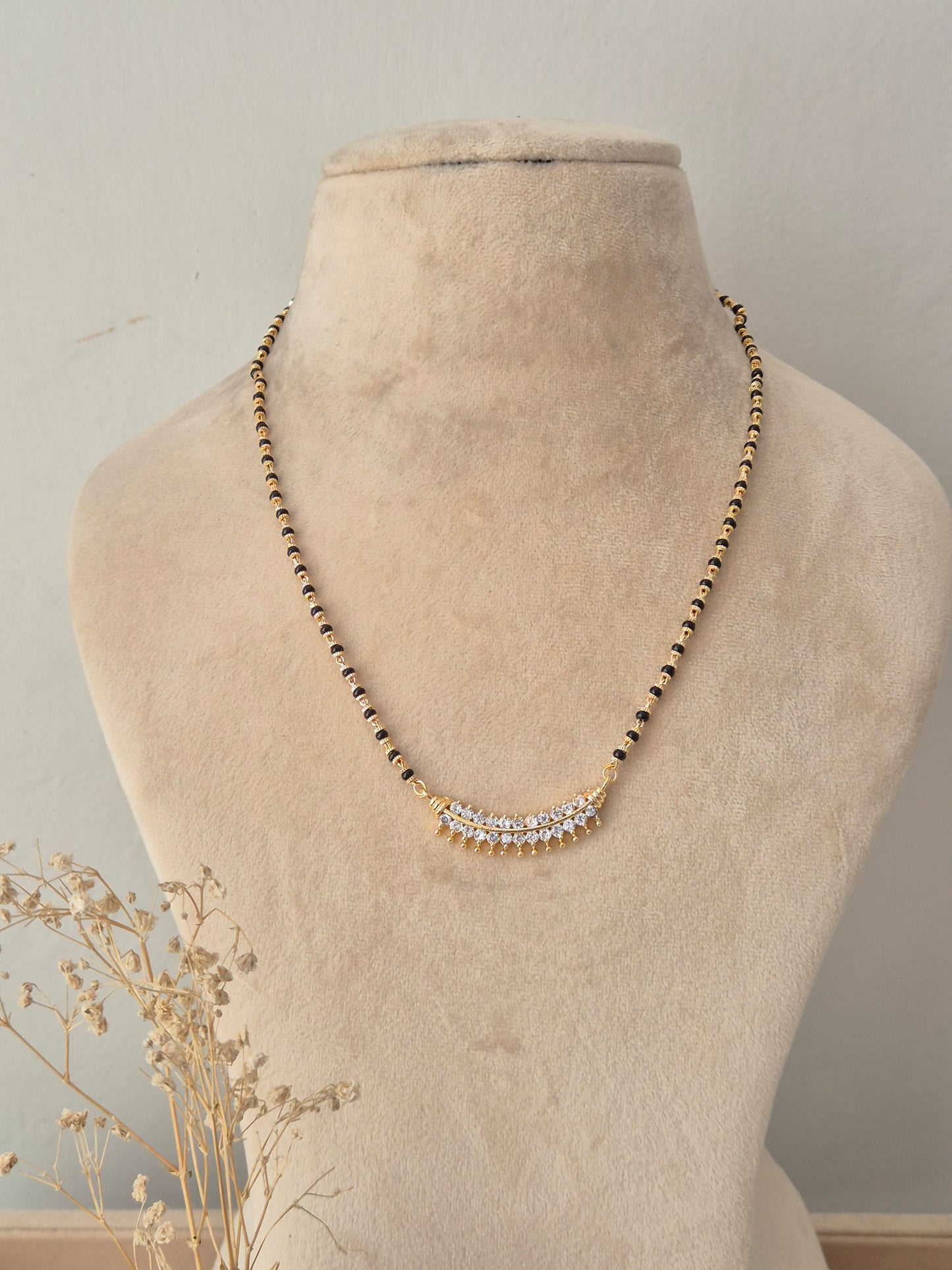 A gold beaded necklace with a curved crystal-embellished pendant, displayed on a beige fabric mannequin bust.