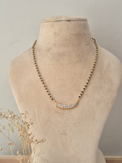 A gold beaded necklace with a curved crystal-embellished pendant, displayed on a beige fabric mannequin bust.