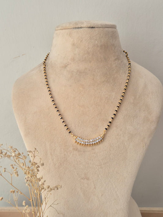 A gold beaded necklace with a curved crystal-embellished pendant, displayed on a beige fabric mannequin bust.