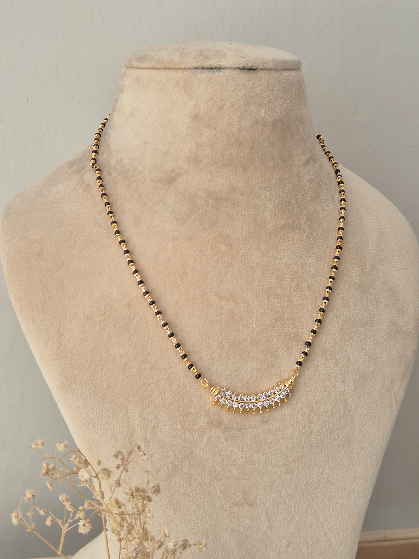A gold beaded necklace with a curved crystal-embellished pendant, displayed on a beige velvet jewelry bust.