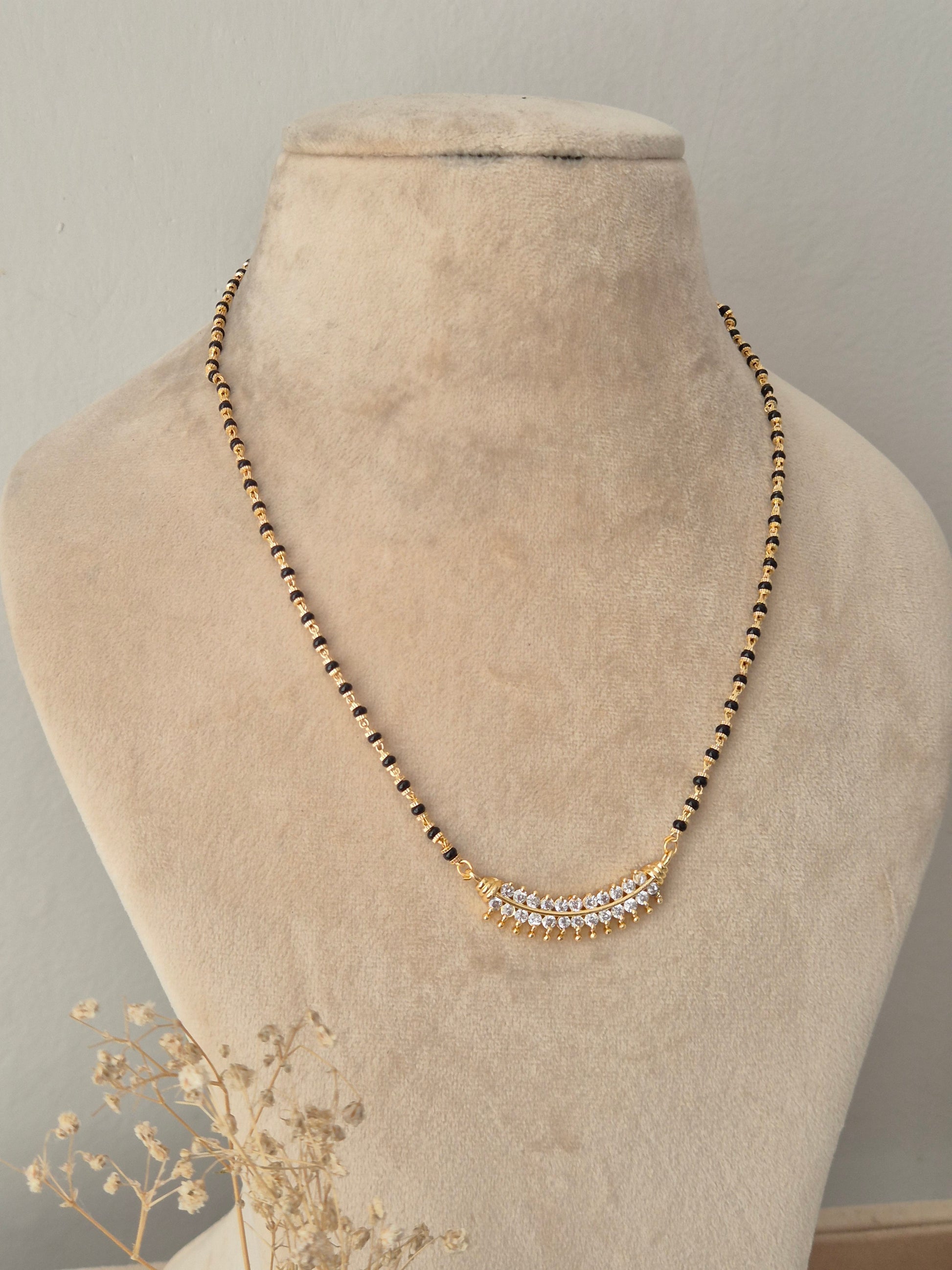 A gold beaded necklace with a curved crystal-embellished pendant, displayed on a beige velvet jewelry bust.