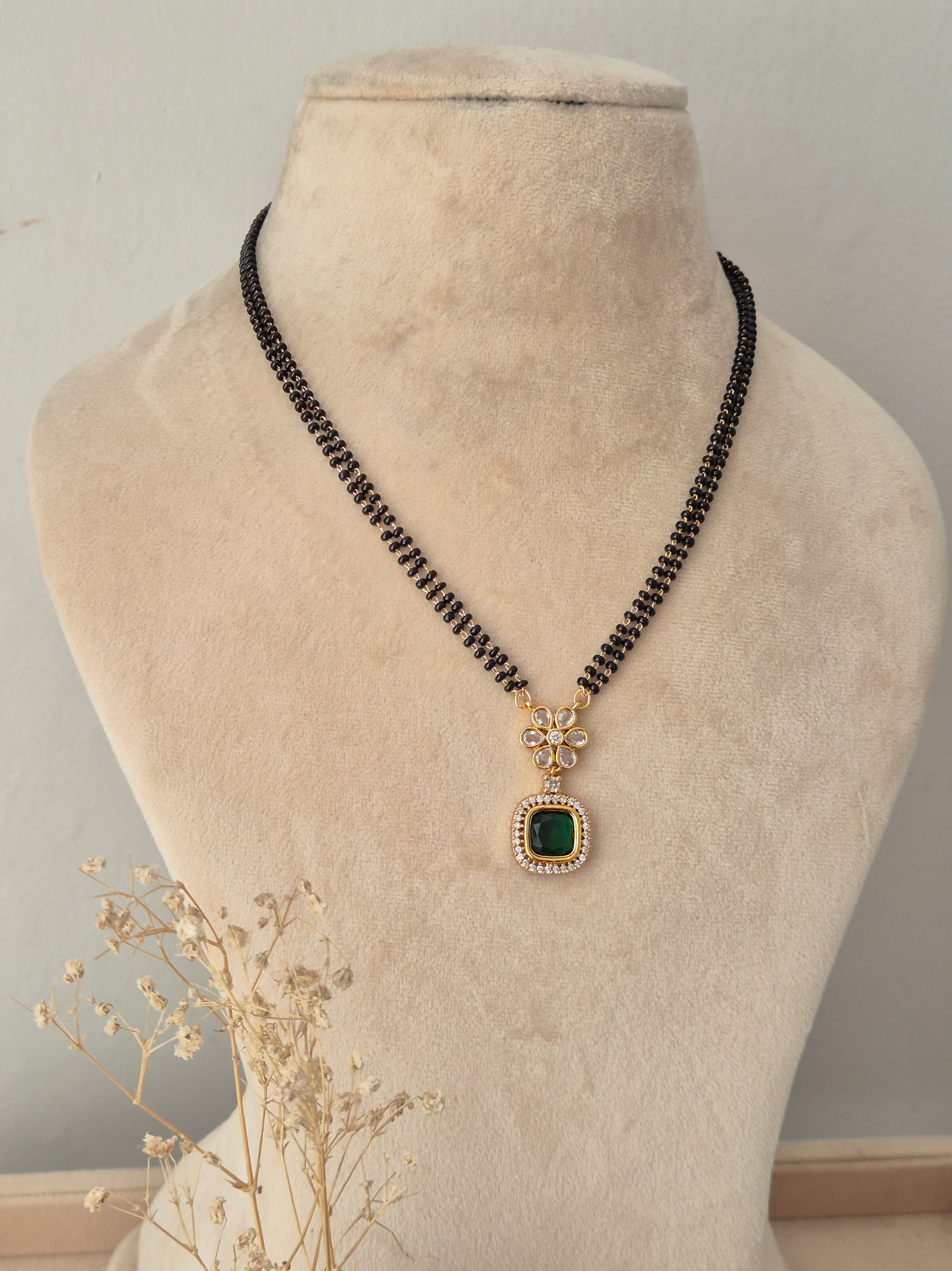 Saloni Mangalsutra featuring double-stranded black beads, a gold floral accent set with clear stones, and a square green stone drop pendant