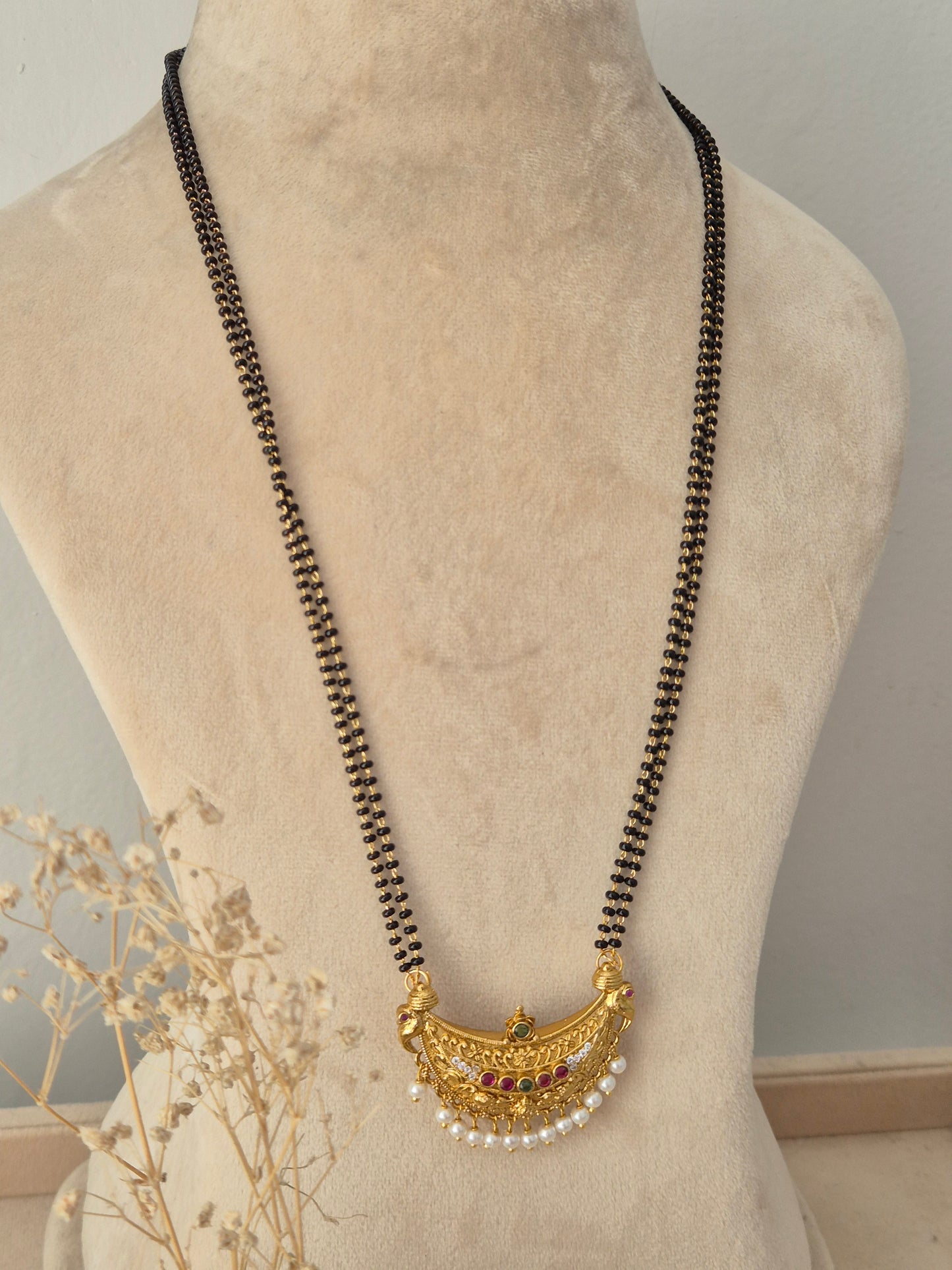 Janani Mangalsutra featuring a long double-strand black bead chain connected to an ornate crescent-shaped gold pendant detailed with red and green stone accents and hanging white pearl beads