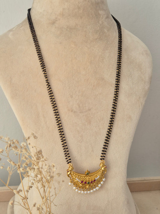 Janani Mangalsutra featuring a long double-strand black bead chain connected to an ornate crescent-shaped gold pendant detailed with red and green stone accents and hanging white pearl beads